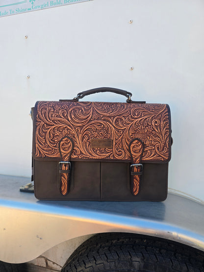 A dark brown leather briefcase with an intricate, tooled floral pattern on the front flap, resting on the silver metal bed of a vehicle against a white trailer wall.