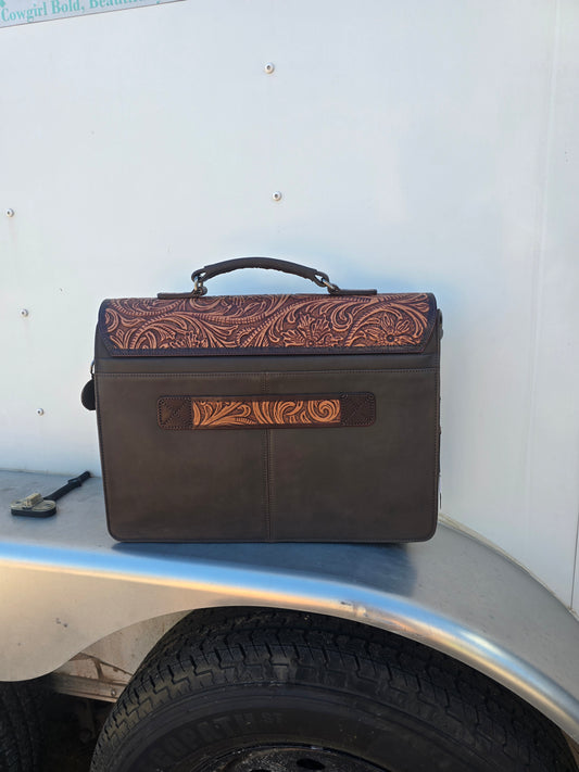 A dark brown leather tool box with tooled brown leather detailing on the top and front, featuring a top handle, placed on the edge of a trailer next to a tire.